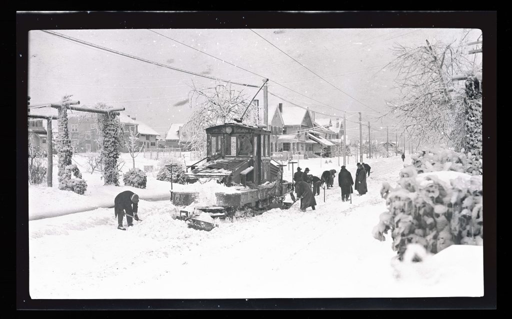 Photograph showing a group of people with a streetcar after a snowfall. The people are using tools and shovels to clear snow around the streetcar train, which is attached to an electric line above. On the side of the train is “P.R.L & P. Co.” Ice laden electric lines can be seen above and around the train, with buildings visible in the background.