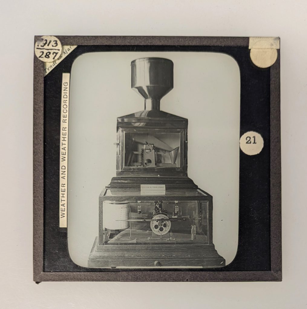A photograph of a lantern slide with several paper labels and showing a photograph of an instrument with a collecting funnel at the top, mechanism in the center and recording apparatus at the bottom.